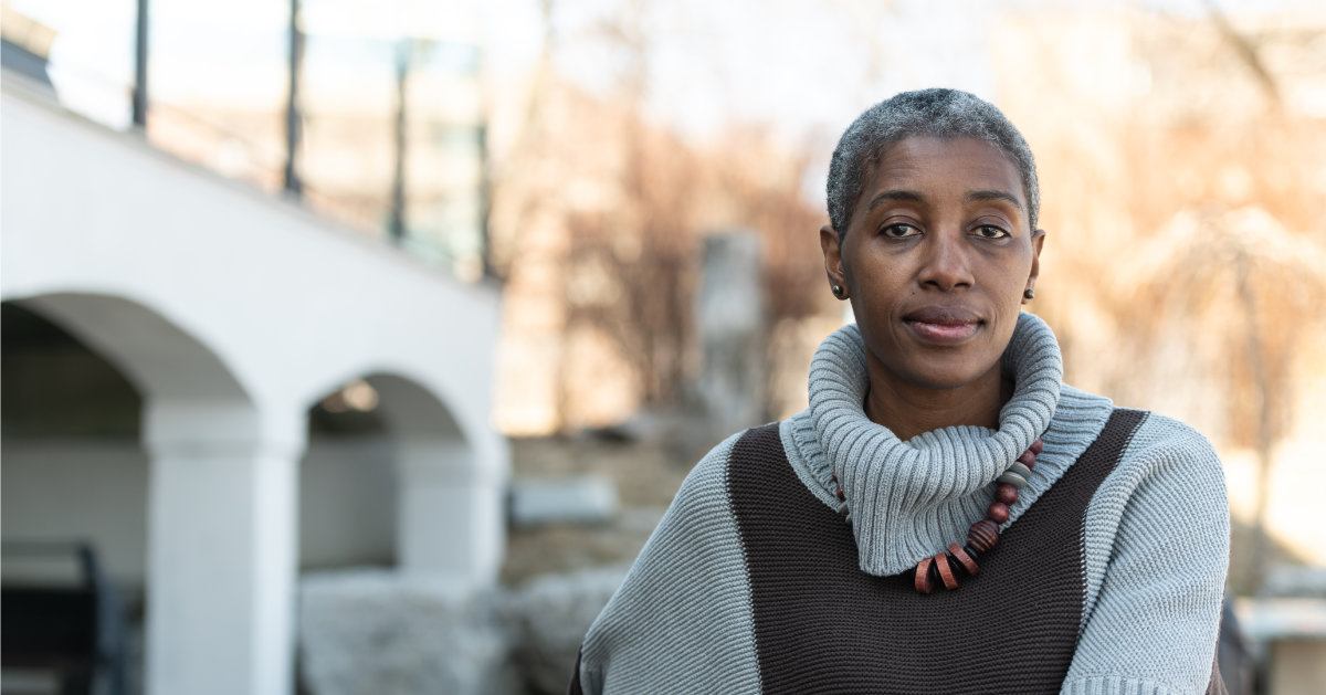 A senior woman of African descent sits outside while looking contemplative. She is dressed warmly in her sweater and looks cozy. She is looking into the camera.