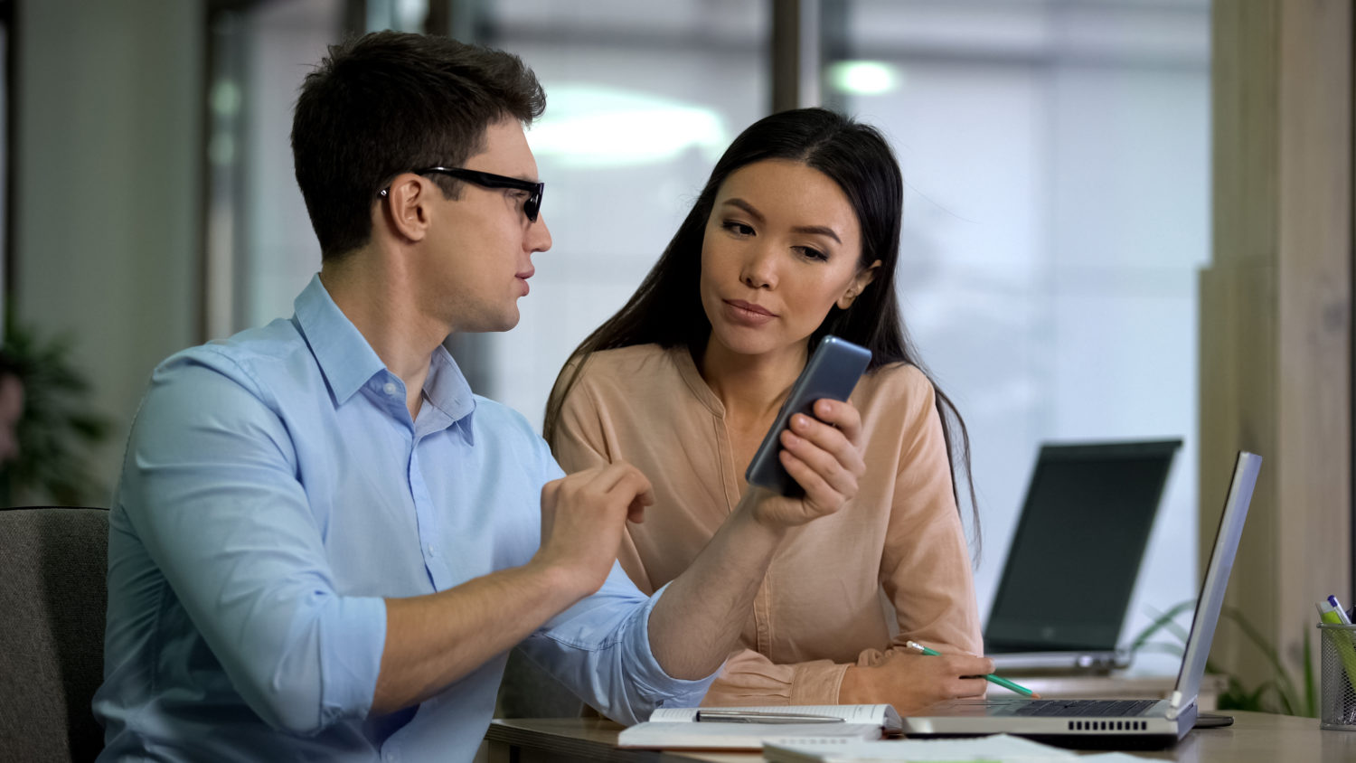 a male colleague shows a female coworker his phone while sitting next to eachother at a desk with a laptop in front