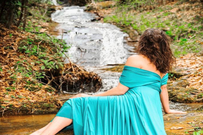 Brunette woman in teal dress facing away from the camera towards a small waterfall