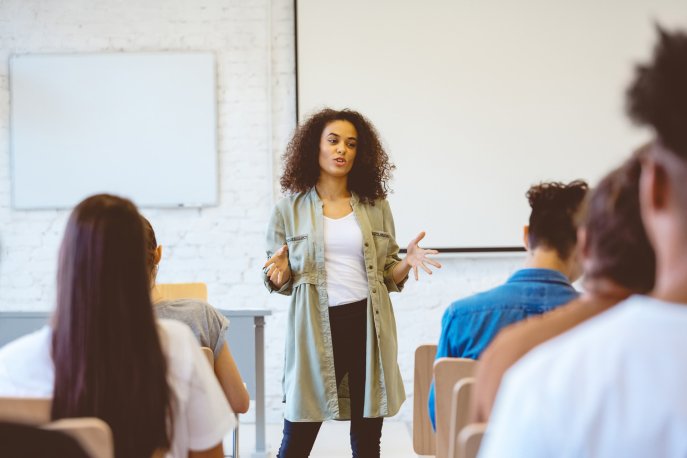 Woman with light brown complexion and brown, curly hair is dressed in a white shirt, black pants, and long green cardigan. She's in the front of a room, facing the camera, running a meeting.