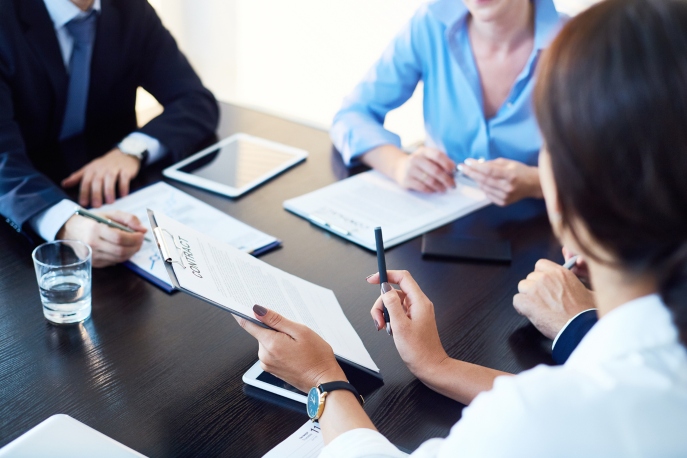 Three people in business attire, one man and two women, seated at a business table overlooking a contract. No faces are visible.