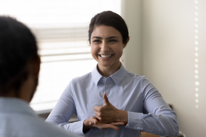 Woman in work blouse smiling and signing the word "help" in ASL.