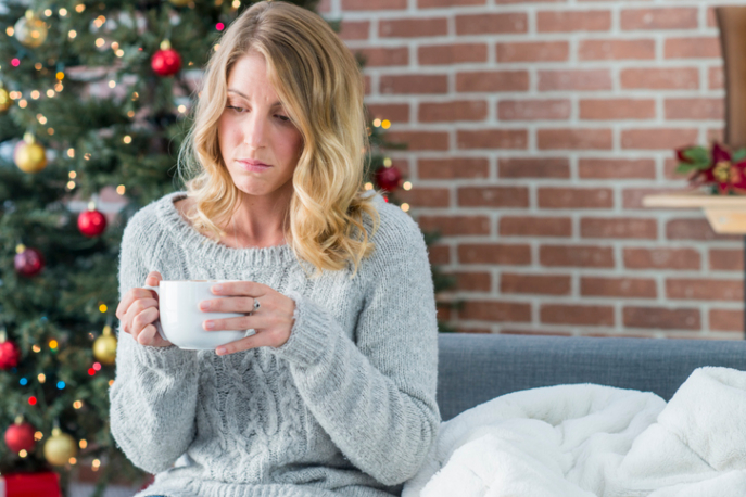Blonde woman seated in front of a Christmas tree holding a mug and looking somber.