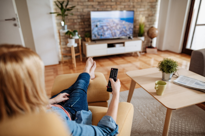 Blonde woman facing away from camera towards tv on her lounge chair, with remote extended in her hand.