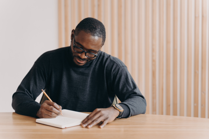 Black man seated at wooden table writing in a journal.