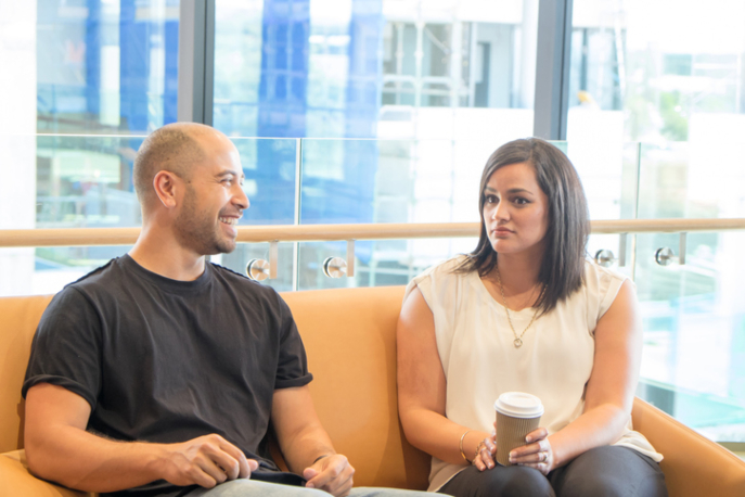 Man and woman seated on a couch. The man is smiling at her while the woman looks away, visibly uncomfortable.