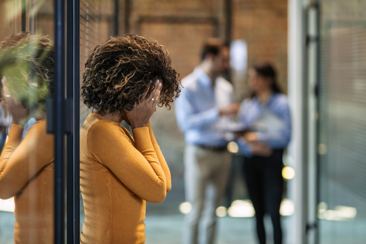 Black woman in yellow long sleeve shirt, leaning against a glass wall at work with her face in her hands.