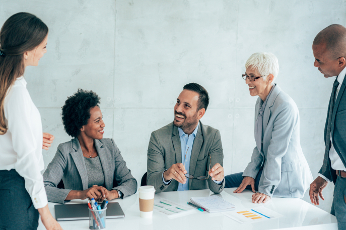 5 employees of various races, ages, and genders smiling and chatting at a conference table.