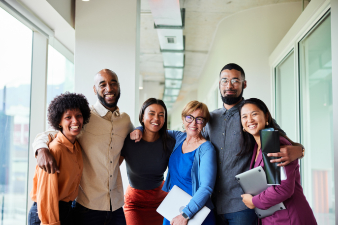 Six employees of varying races and genders, arms around each other, facing the camera and smiling.