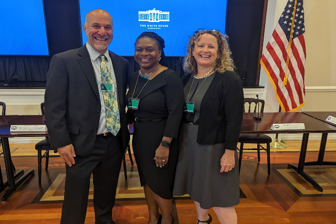 David Lee, a white man, Monika Johnson-Hostler, a Black woman, and Terri Poore, a white woman, smiling together in a conference room with a screen featuring the White House logo in the background.