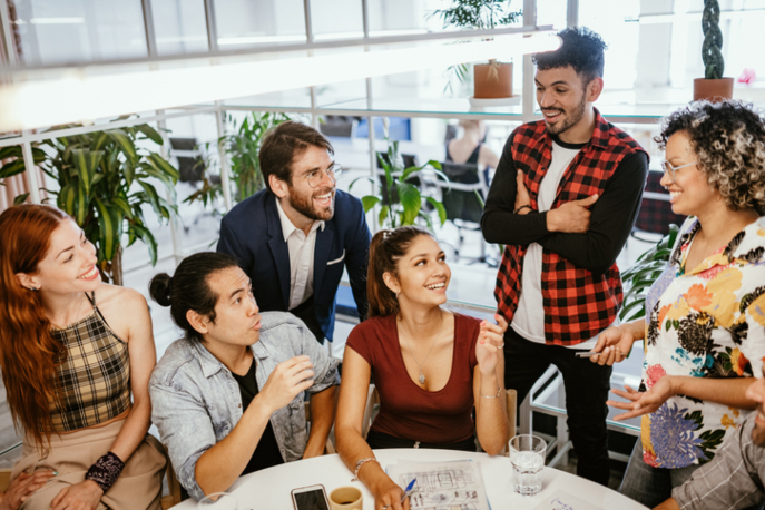 Group of six adults of varying races and genders, standing and sitting in front of a table, smiling.