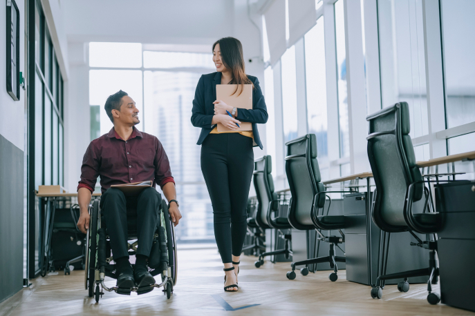 Latino man in wheelchair traveling with able-bodied Asian woman down business hallway.