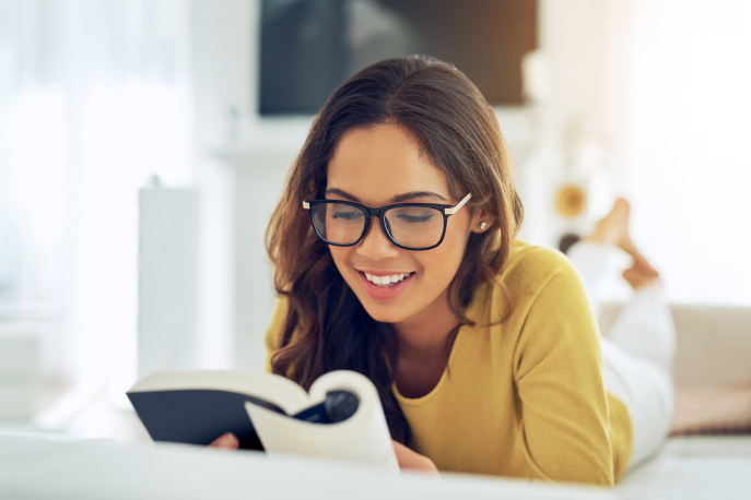 Tan brunette woman in glasses and a yellow sweater lying on her stomach, reading a book happily.