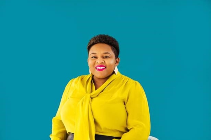 Black woman with short hair and a yellow blouse on a teal background.