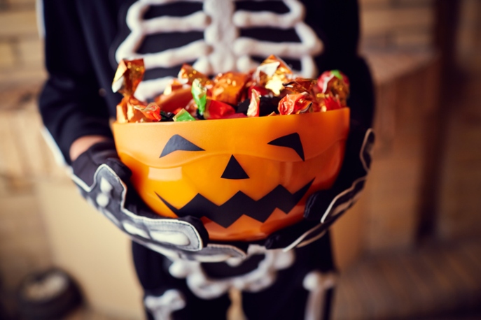 Pumpkin basket of halloween candy held by child in skeleton costume. View of child is shoulder to leg.