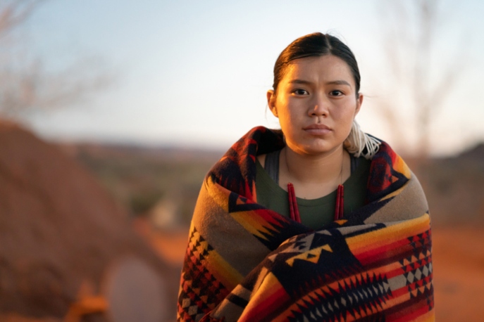 Native American woman, wrapped in a blanket, staring at the camera somberly.