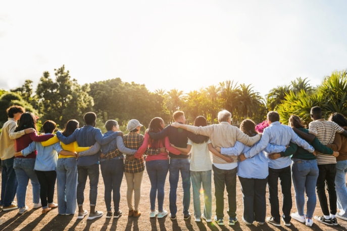 Adults of various races with their arms around each other, facing away from the camera and towards a sunset