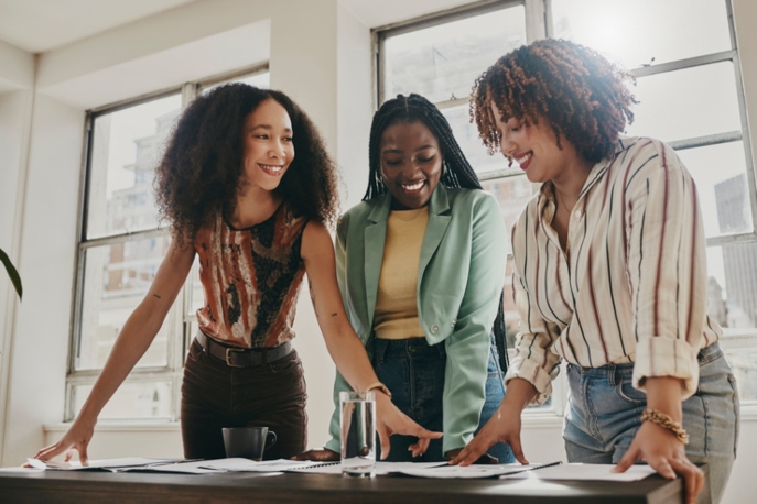 Three professional Black women smiling at each other, in front of a table of documents.