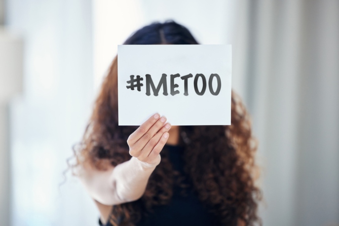 Curly haired brunette woman holding a #MeToo sign in front of her face.