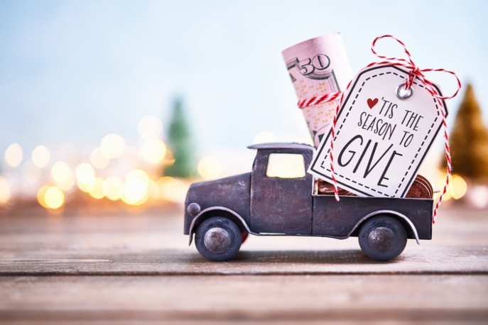 Truck carrying roll of dollars with holiday background. A sign says, "Tis the season to give"