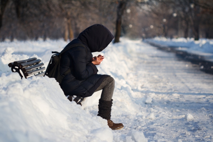 Person in a hoodie and backpack staying on a bench on a snowy street.